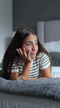 Pensive young woman resting on a cozy bed in a dim bedroom, looking away pensively while daydreaming, sighing, and reflecting on thoughts during nighttime relaxation at home