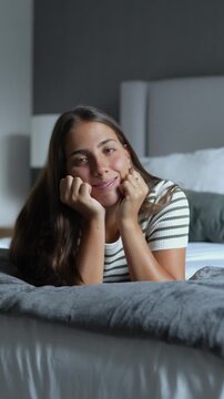 Beautiful young woman lying on a comfortable bed with head resting on hands, smiling, daydreaming and looking thoughtfully at the camera in a cozy bedroom