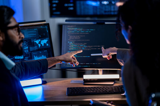 Two coworkers pointing at the same line of source code together, his finger and her stylus almost meeting on the monitor during an evening software debugging session