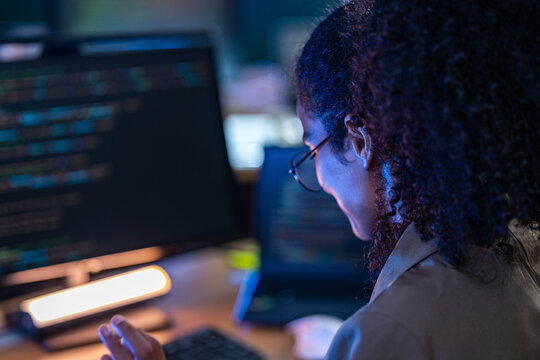 Over-the-shoulder shot of a smiling woman in glasses typing on a keyboard beside a large monitor full of colorful source code, coding late at night at her wooden desk