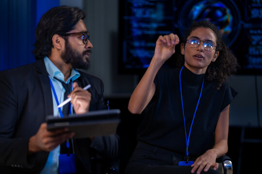 Curly haired woman touches her hair and looks up while thinking aloud as her colleague turns toward her with a tablet and stylus, lines of code glowing on a screen behind