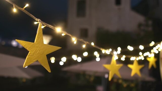 Garland hung on a tree in a garden in the evening and yellow wooden decorative stars. Fashion decoration with bulbs for night summer party. Outdoor electric lamps
