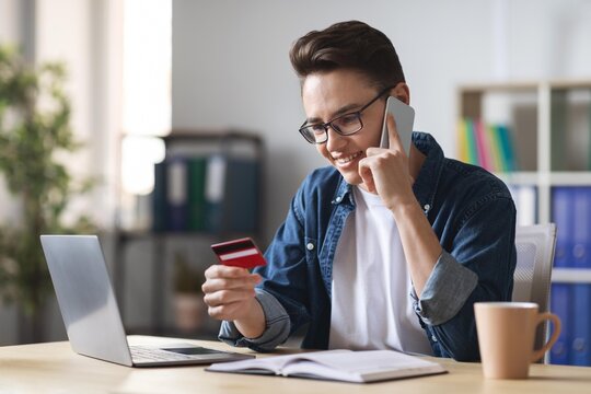 Smiling Young Caucasian Man Holding Credit Card While Talking On Smartphone, Placing Order Online At Home Office Desk. Happy Emotion In Professional Environment For Business Concept.