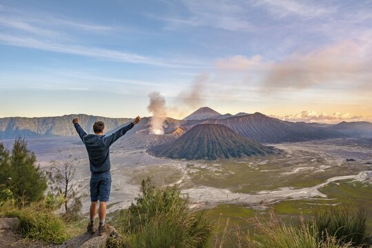 Young man stretches arms in the air, volcanic landscape at sunset, view in Tengger Caldera, smoking volcano Gunung Bromo, in front Mt. Batok, in the back Mt. Kursi, Mt. Gunung Semeru, National Park Bromo-Tengger-Semeru, Java, Indonesia