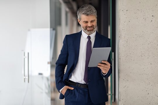 Focused Caucasian Man In Suit Smiling While Looking At Tablet In Modern Office Setting. Engaged In Work Activity, Experiencing Professional Success In Corporate Environment.