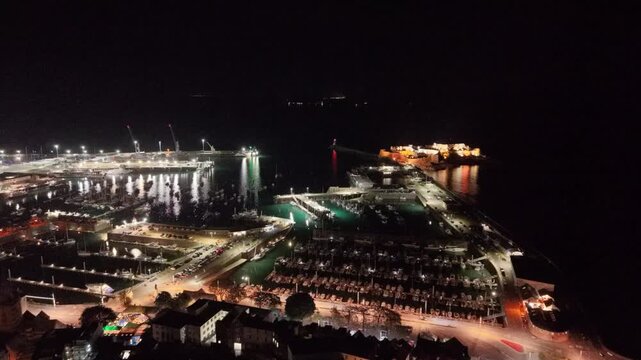 St Peter Port Harbour.Guernsey,Channel Islands.Slow high forward flight at night from seafront towards harbour entrance,docks,ferry berth and Castle Cornet with bright lights reflecting in water