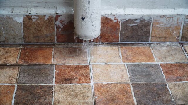 Static tripod shot of water dripping from a weathered drain pipe onto a tiled floor, creating a small wet area. Useful for plumbing, water leakage, household infrastructure, and conservation