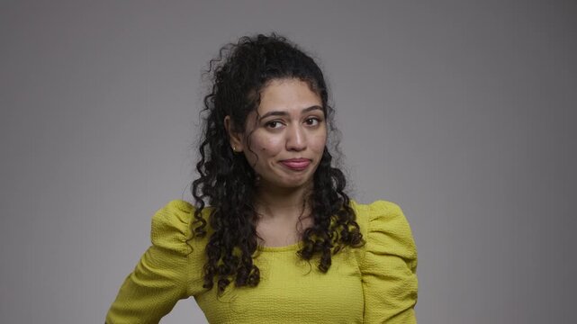 Woman in a yellow top rests a hand at her hip and shifts her gaze sideways with pursed lips and a skeptical look.
