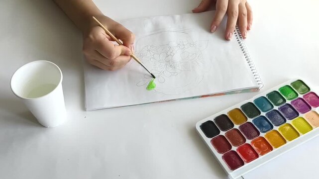 Teenage girl painting in a sketchbook with a watercolor brush, carefully adding color to her drawing, illustrating creativity, artistic development, self-expression, and learning through art.