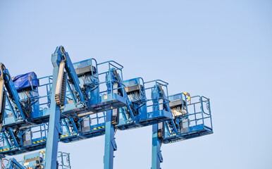 Blue boom lift baskets viewed from below against a clear sky, with guardrails, control boxes and hydraulic hoses visible in a close composition leaving copy space above and to the right in daylight © Fahroni