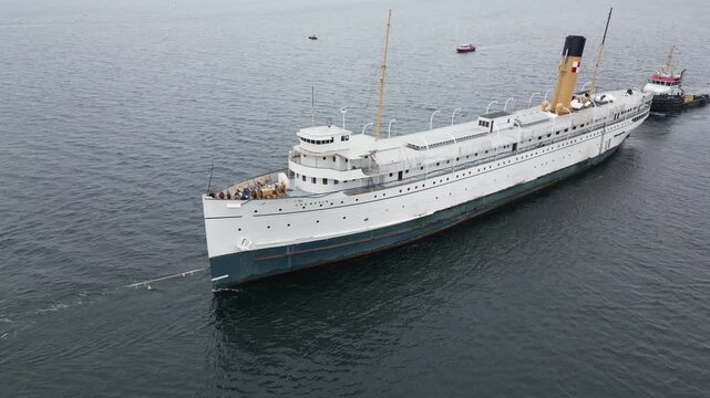 Drone view of a historic white steamship with a yellow funnel being towed across calm grey water, small boats nearby.