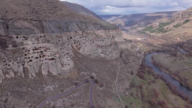Panoramic drone flight follows the Mtkvari River valley before revealing the magnificent Vardzia Cave Town carved into towering cliffs in southern Georgia.