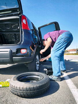 Man bent over loosening lug nuts on alloy wheel with tubular wrench. Conveys vehicle maintenance cost savings, practical car repair skills, and preparedness for unexpected roadside emergencies.