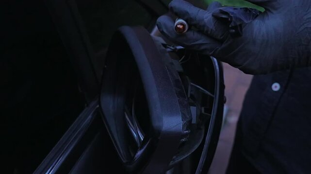 Gloved hands replace a small turn signal bulb inside a damaged car side mirror housing at dusk.