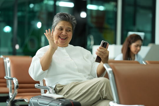 A mature female traveller in a white blouse holds her passport and boarding pass while she waits on the seats of an airport gate area, with her wheeled case beside her