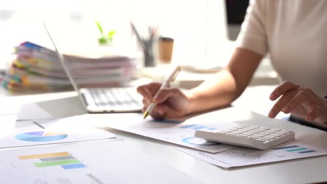 Financial Analysis at Desk: A focused individual meticulously analyzes financial reports and charts with a calculator and laptop at a bright, organized workspace, symbolizing diligence and planning.