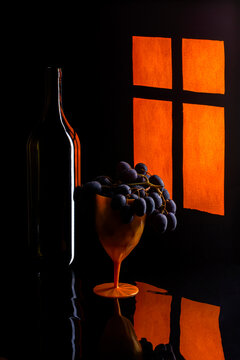 Still life with a bunch of grapes and a bottle near a lit window.