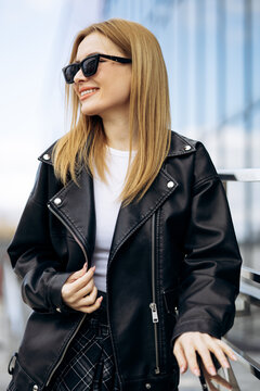 Young blonde woman smiling stylishly in sunglasses, wearing a black leather jacket and checkered skirt in an urban setting