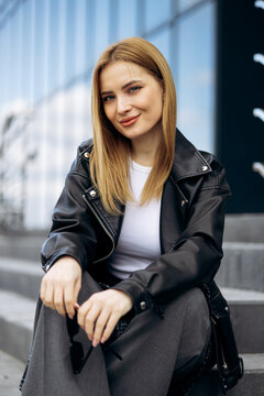 Blonde woman sitting on urban steps, holding sunglasses and posing, exuding confidence and casual style