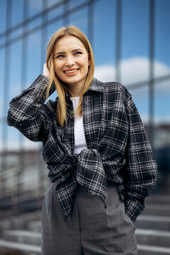 Young blonde woman posing outdoors, wearing a long sleeve plaid shirt and grey trousers against a modern glass building