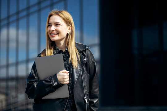 Young blonde woman smiling, holding a laptop in hand, standing confidently in front of a modern urban building