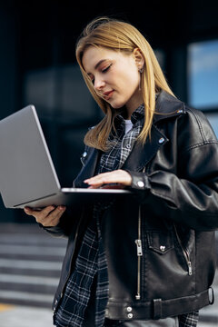 Young woman dressed in a stylish leather jacket and plaid shirt, typing on a laptop computer while standing outside