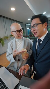 Two professionals share a joyful moment at their desk. One sips coffee while the other laughs heartily. They wave cheerfully, radiating team spirit