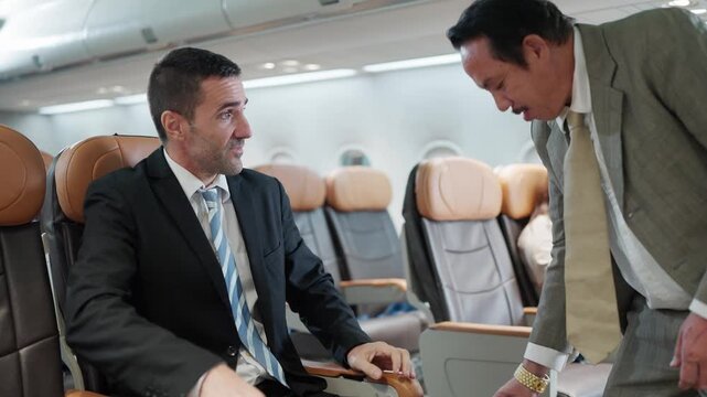 Two businessmen on an airplane. One man stands, pulling a suitcase. Another man sits, watching. They smile, sharing a moment. Professional travel