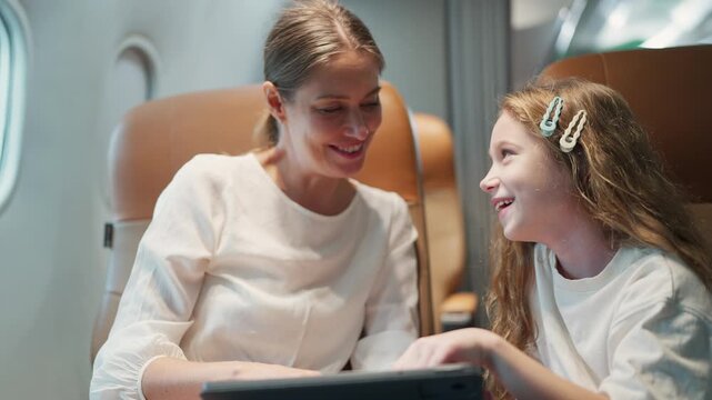 Mother and daughter laugh together on a flight. They share a tablet, smiling and looking at each other. Soft lighting shows comfort