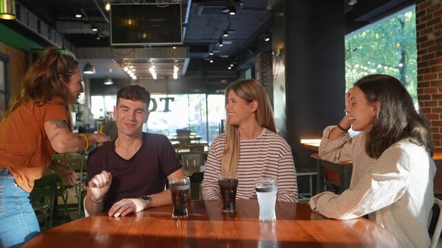 Three young friends are enjoying their time together, talking and laughing at a table, when a fourth friend arrives, greeting them with warm hugs before she finally leaves