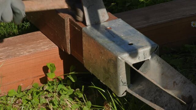 Close-up of a worker's gloved hand using a hammer to assemble a brown wooden structure with galvanized steel brackets in a garden