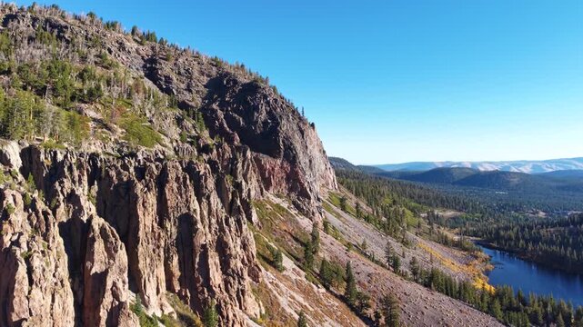 Rugged rock cliffs above a forested valley and blue mountain lake under clear skies, aerial pullback along steep sharp drop off sloping down to water
