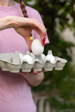Woman's hand with red painted nails carefully selecting a fresh white chicken egg from a cardboard carton, emphasizing concepts of natural food, organic ingredients, and healthy eating habits