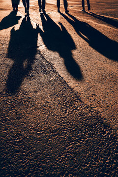 Long dark shadows of a group of people walking across warm orange asphalt at sunset, with only their legs visible at the top and a rough textured ground surface in the foreground.