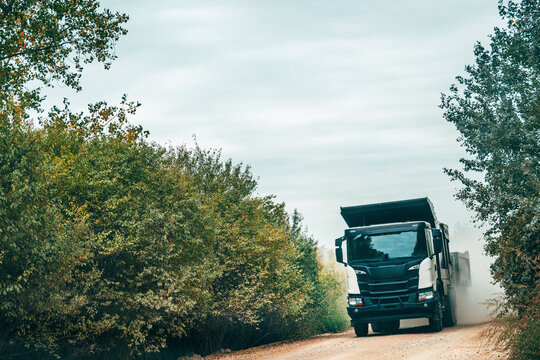 Heavy dump truck driving toward the camera on a dusty unpaved road between green and yellow autumn trees, with a raised cargo bed, rising dust and a hazy overcast sky above.