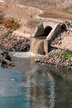 Brown wastewater flowing from a concrete drainage outlet into a river along a stone embankment, with rubble and a fallen log, environmental pollution concept
