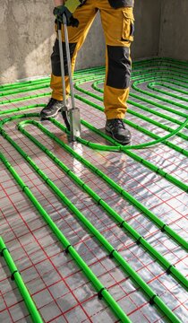 Worker installing green underfloor heating pipes on a foil insulation layer with a red grid using a clip tool, radiant floor heating system in a new building