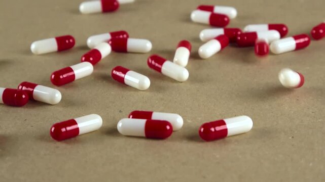 Red and white medical capsules falling continuously onto a flat brown cardboard surface, bouncing and scattering in slow motion during a pharmaceutical concept shoot