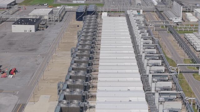 Long rows of air cooled chiller units with stacked fans line both sides of a white roofed data center hall, with blue water tanks, pipe racks and an electrical substation beyond. Aerial view