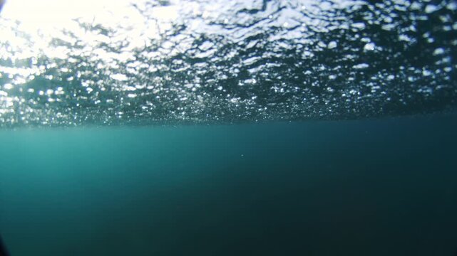 Midday sunlight glistens through rippled ocean surface above deep teal water, underwater sparkling abstract texture