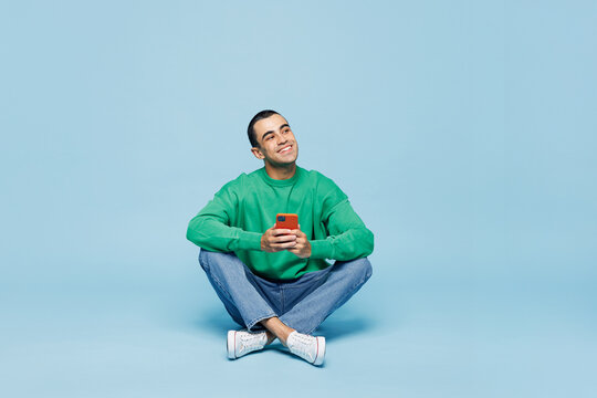 Full body young happy man wear shirt white t-shirt casual clothes sit hold in hand use mobile cell phone look aside on area isolated on plain blue cyan background studio portrait. Lifestyle concept