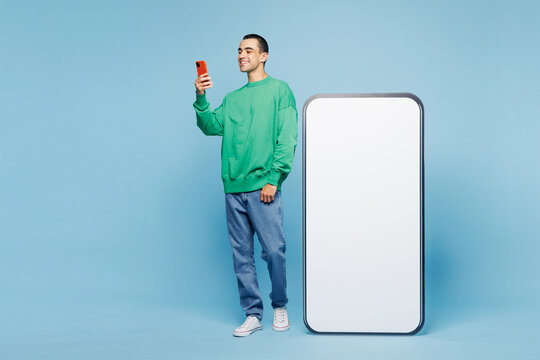 Full body young fun middle eastern man wear green shirt casual clothes stand near big huge blank screen mobile cell phone with copy space mock up area use smartphone isolated on plain blue background.