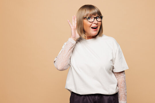 Elderly curious nosy blonde woman 50s years old wear white shirt casual clothes try to hear you overhear listening intently isolated on plain pastel light beige background studio. Lifestyle concept.