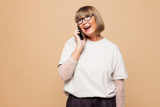 Elderly blonde woman 50s years old wear white shirt casual clothes talk speak on mobile cell phone conducting pleasant conversation isolated on plain pastel light beige background. Lifestyle concept.