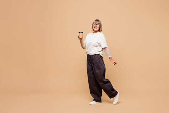 Full body elderly blonde woman 50s years old she wear white shirt casual clothes hold takeaway delivery craft paper brown cup coffee to go isolated on plain pastel beige background. Lifestyle concept.