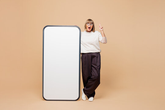 Full body elderly woman 50 years old wear white shirt casual clothes big huge blank screen mobile cell phone smartphone point finger up with area isolated on plain beige background. Lifestyle concept