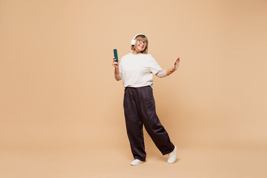 Full body happy elderly blonde woman 50s years old she wear white shirt casual clothes listen to music in headphones use mobile cell phone isolated on plain pastel beige background. Lifestyle concept.