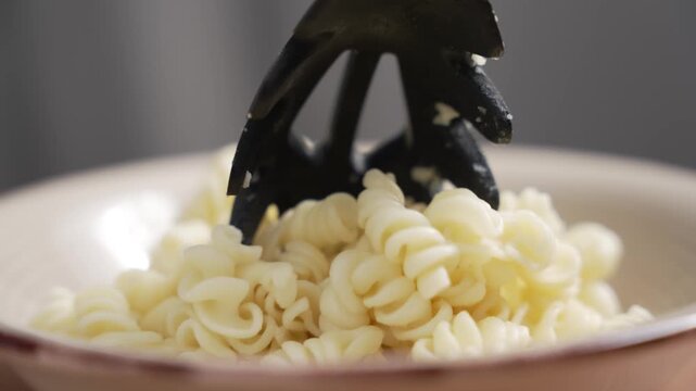 Cooked fusilli pasta is dropped from a black pasta server onto a pile of plain spiral pasta in a ceramic bowl. Close-up view.
