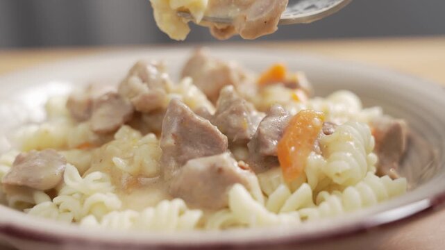 A hand uses a fork to pick up meat chunks and fusilli pasta in creamy sauce from a plate. Close-up with carrot pieces visible.