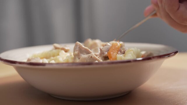 Low side view of a ceramic bowl of fusilli pasta with creamy meat stew and carrot on a table as a hand takes forkfuls of food.